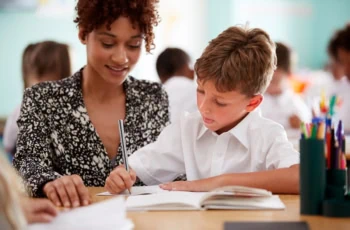 Woman Elementary School Teacher Giving Male Pupil Wearing Uniform One To One Support In Classroom