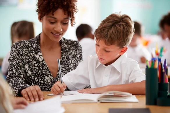 Woman Elementary School Teacher Giving Male Pupil Wearing Uniform One To One Support In Classroom