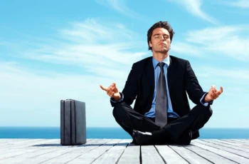 a businessman meditates in a yoga pose while sitting on a dock with his briefcase resting nearby.