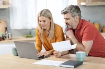 Happy middle aged husband and wife sitting at kitchen table in front of computer, holding papers, working on family budget together, paying bills online