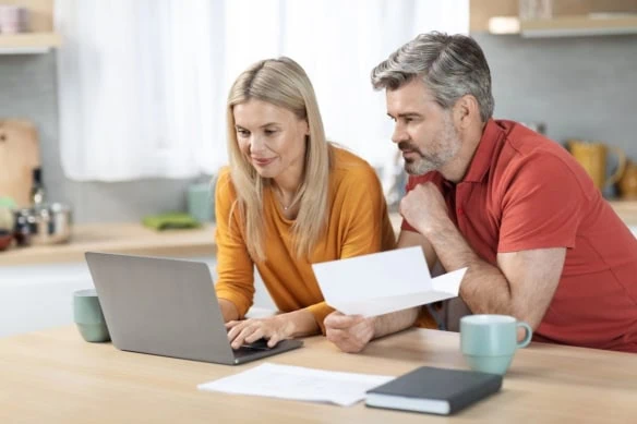 Happy middle aged husband and wife sitting at kitchen table in front of computer, holding papers, working on family budget together, paying bills online