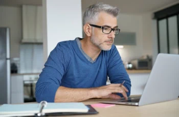 middle aged man looking at laptop with notebook taking notes