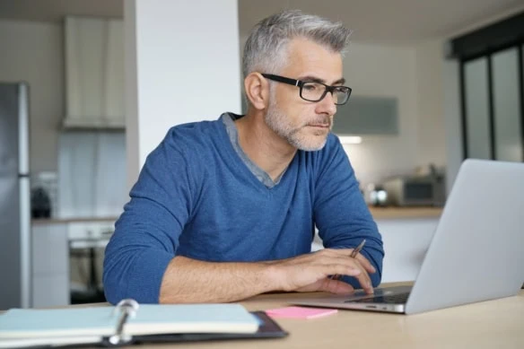 middle aged man looking at laptop with notebook taking notes