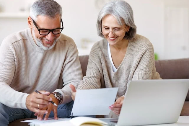 Smiling senior couple reading notification letter with good news from bank while sitting with laptop