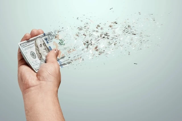 A hand holds a hundred dollar bills that dissolve into thin air. The concept of disappearing money