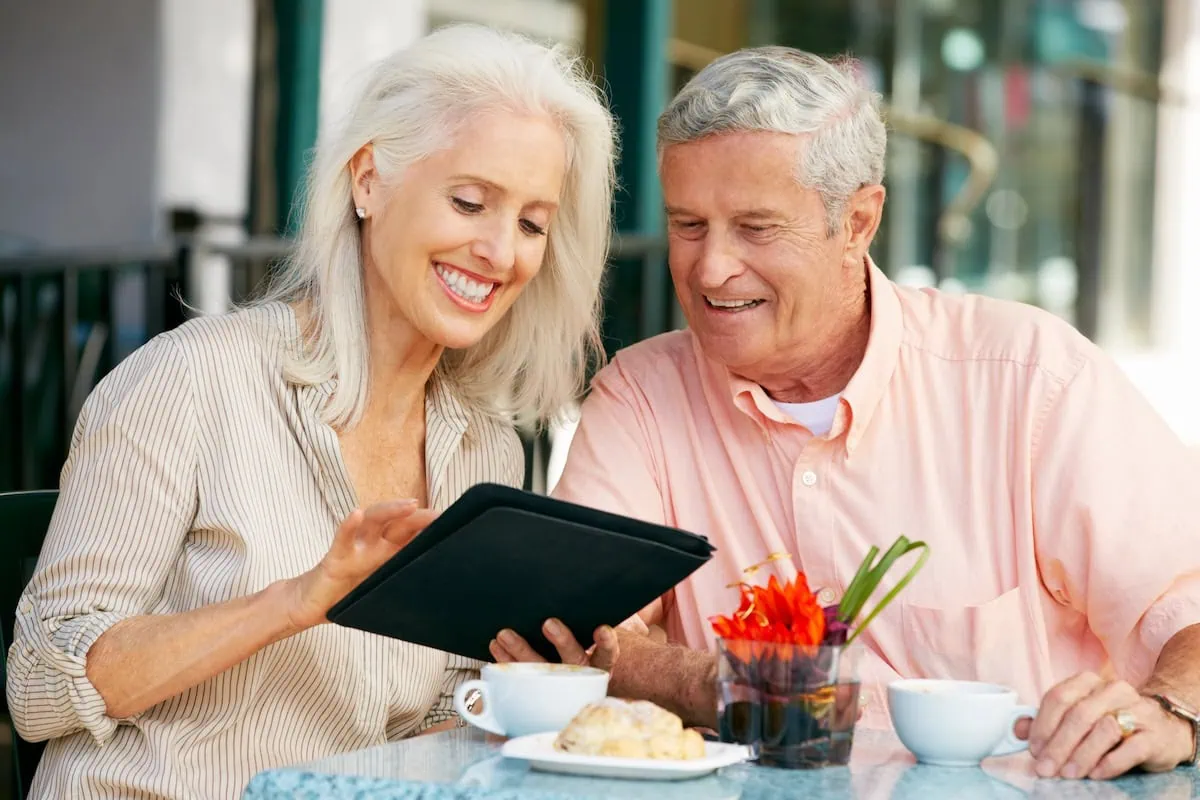 Are You Collecting an Above-Average Check? These Are the Average Monthly Social Security Payments by Age 2 Senior Couple Using Tablet Computer At Outdoor Café