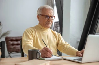Senior man sitting at the table with modern laptop and taking some notes