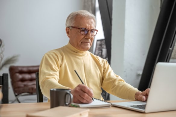 Senior man sitting at the table with modern laptop and taking some notes