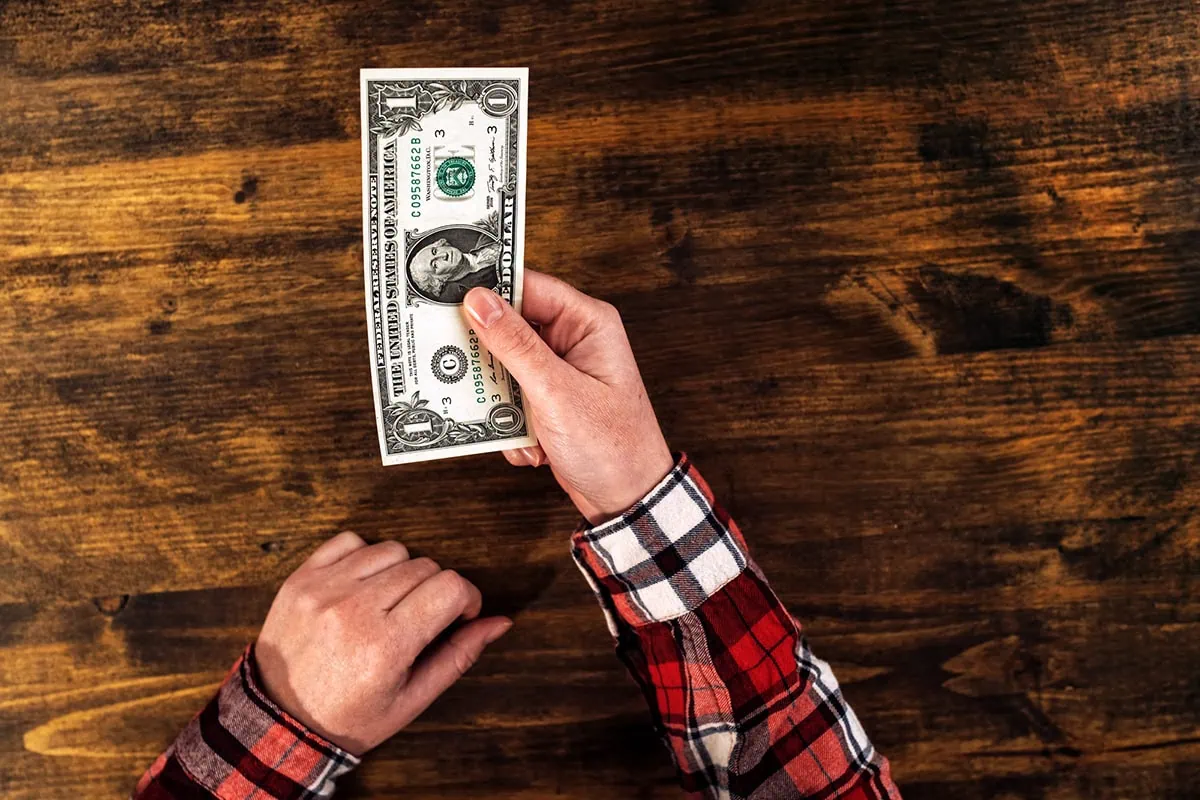 a person in a plaid shirt hands over a one dollar bill.