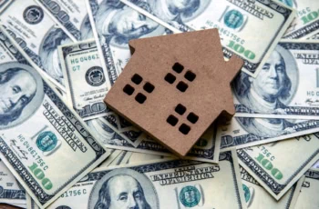 a wooden block model of a home lies on a pile of cash.