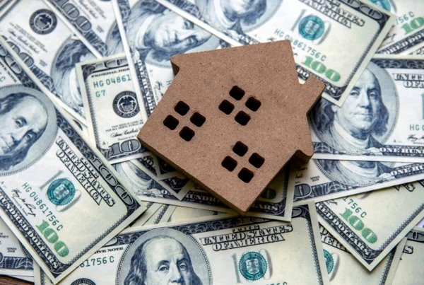 a wooden block model of a home lies on a pile of cash.
