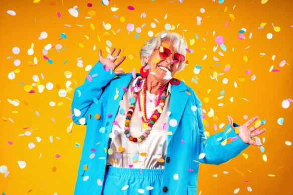 a senior woman wearing an oversized blue suit celebrates around confetti.