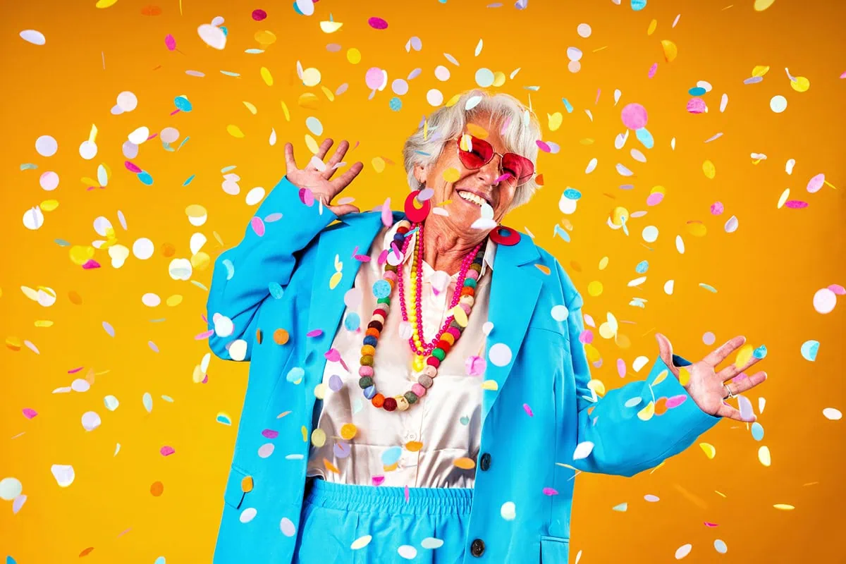 The Seniority Secret: Dozens of Age-Based Discounts That Are Often Overlooked 2 a senior woman wearing an oversized blue suit celebrates around confetti.