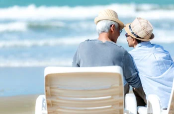 An older Asian couple look at each other while sitting on chairs beside a beach.