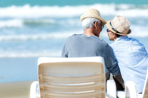 An older Asian couple look at each other while sitting on chairs beside a beach.