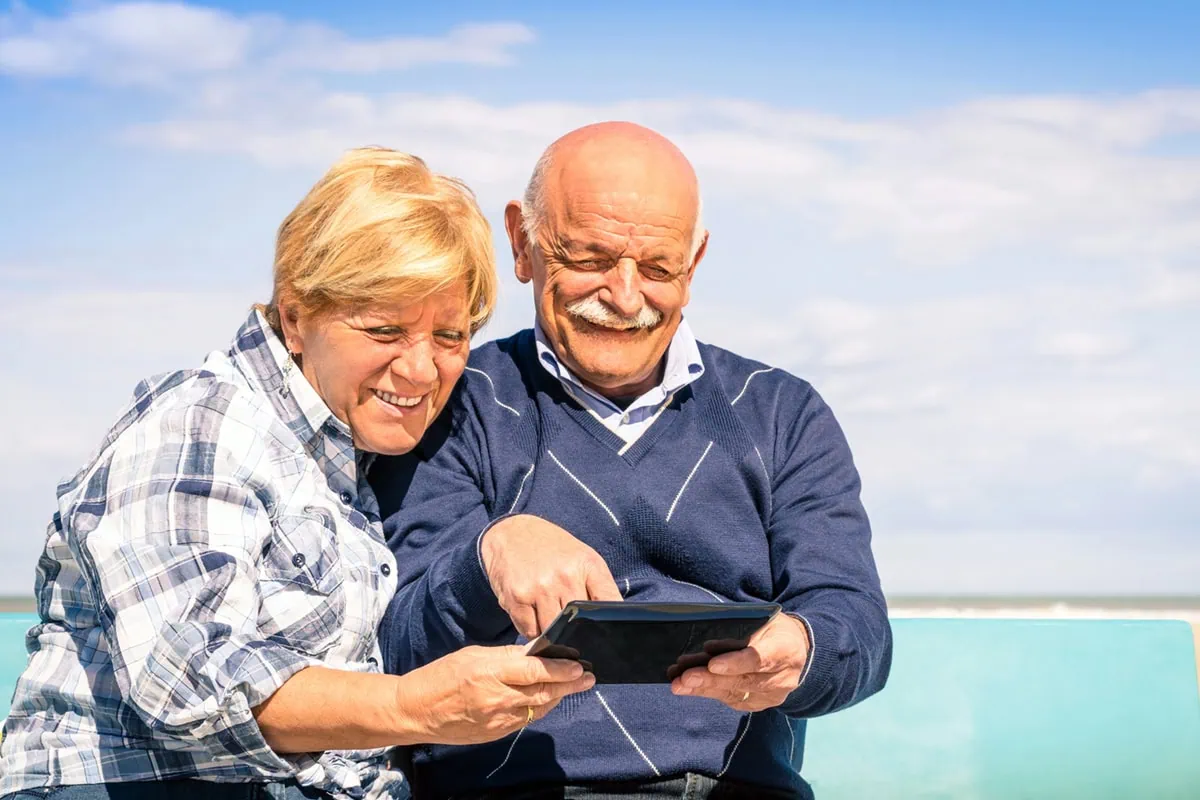 15 Major Companies with Special Discounts for Seniors 2 a couple looks at a tablet while sitting by the beach on a sunny day.