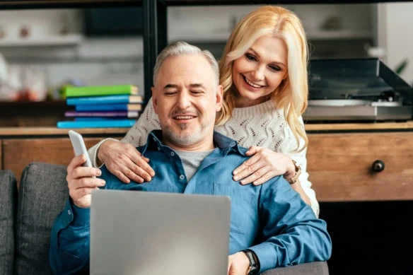 an older couple smiles while looking at their computer.