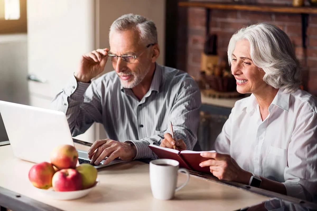 Senior couple sitting at the kitchen table looking at something on a laptop and smiling.