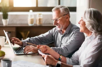 a senior man and woman smile while studying something on their laptop.