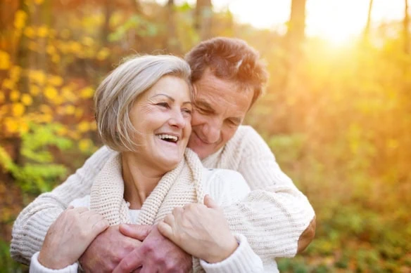 a happy retired couple holds one another in the forest during fall.