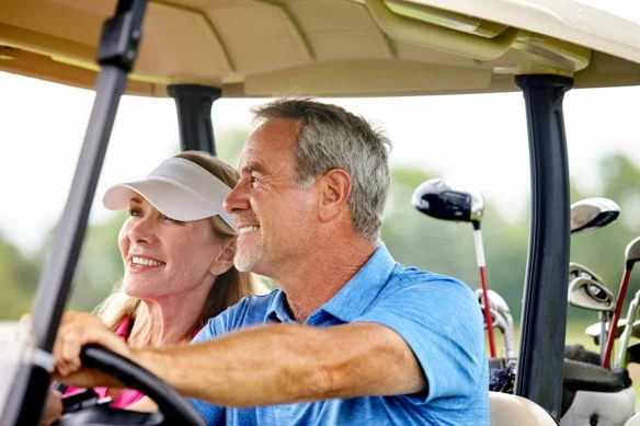 a retired man and woman smile while driving a golf cart.