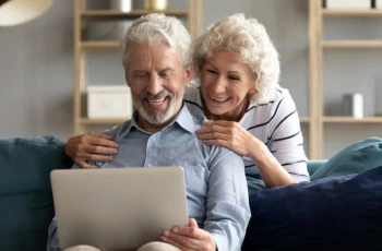an older man and woman smile while looking at their laptop.