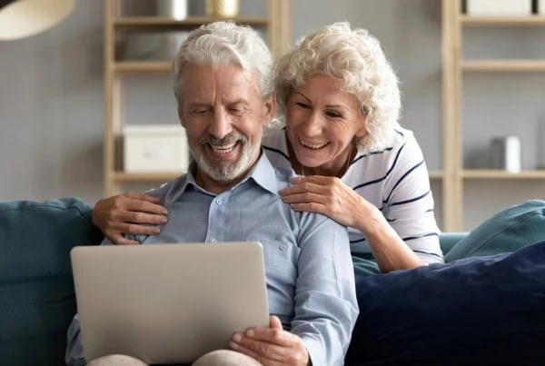 an older man and woman smile while looking at their laptop.