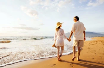 a retired couple walks barefoot on a beach on a sunny day.