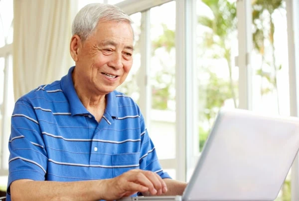 an older Chinese man smiles while looking at his laptop.
