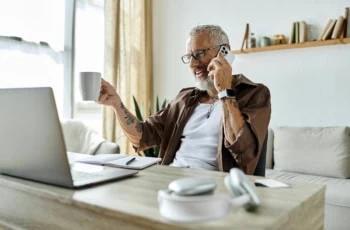 a man with numerous tattoos on his arms drinks coffee and talks on the phone in front of his laptop.