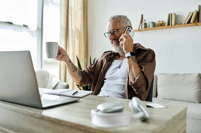 a man with numerous tattoos on his arms drinks coffee and talks on the phone in front of his laptop.