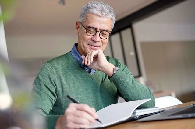 an older man wearing a green shirt looks at his finances on his computer and a notebook.