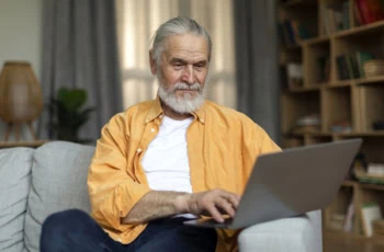 an older man in an orange shirt types on his laptop while sitting on a couch.