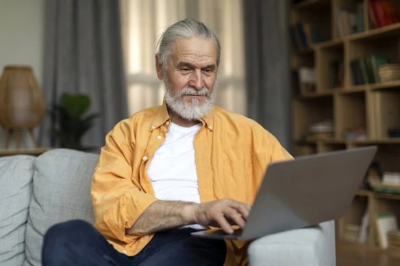 an older man in an orange shirt types on his laptop while sitting on a couch.