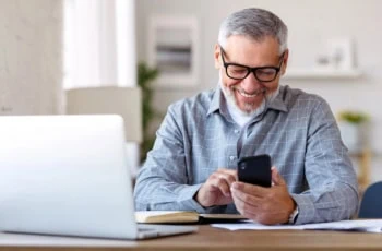 a man smiles while looking down at his smartphone at his desk at home.