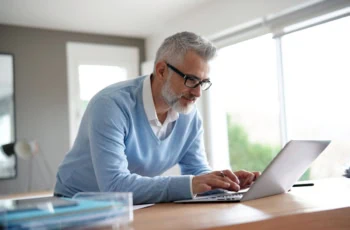 a middle aged man in a blue sweater and white button up uses his laptop in an office.