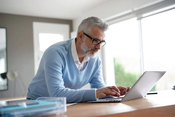 a middle aged man in a blue sweater and white button up uses his laptop in an office.