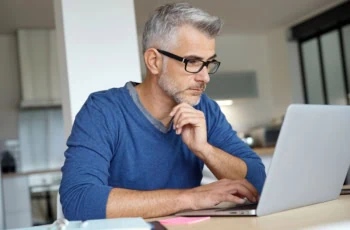 a middle-aged man wearing a blue shirt looks at his laptop while working in his home kitchen.