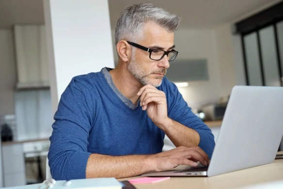 a middle-aged man wearing a blue shirt looks at his laptop while working in his home kitchen.
