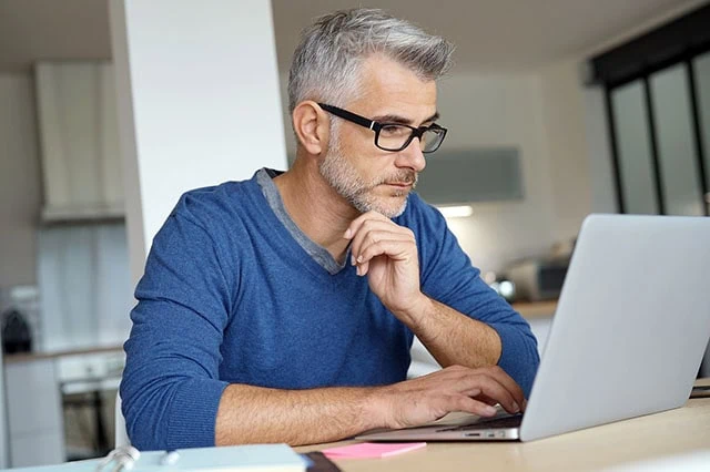 a middle-aged man wearing a blue shirt looks at his laptop while working in his home kitchen.