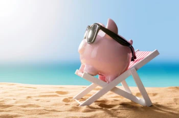 a piggy bank wears sunglasses and sits on a beach chair resting on the sand.