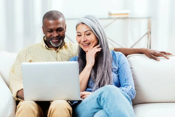 a happy senior couple looks at a computer while sitting at their couch.