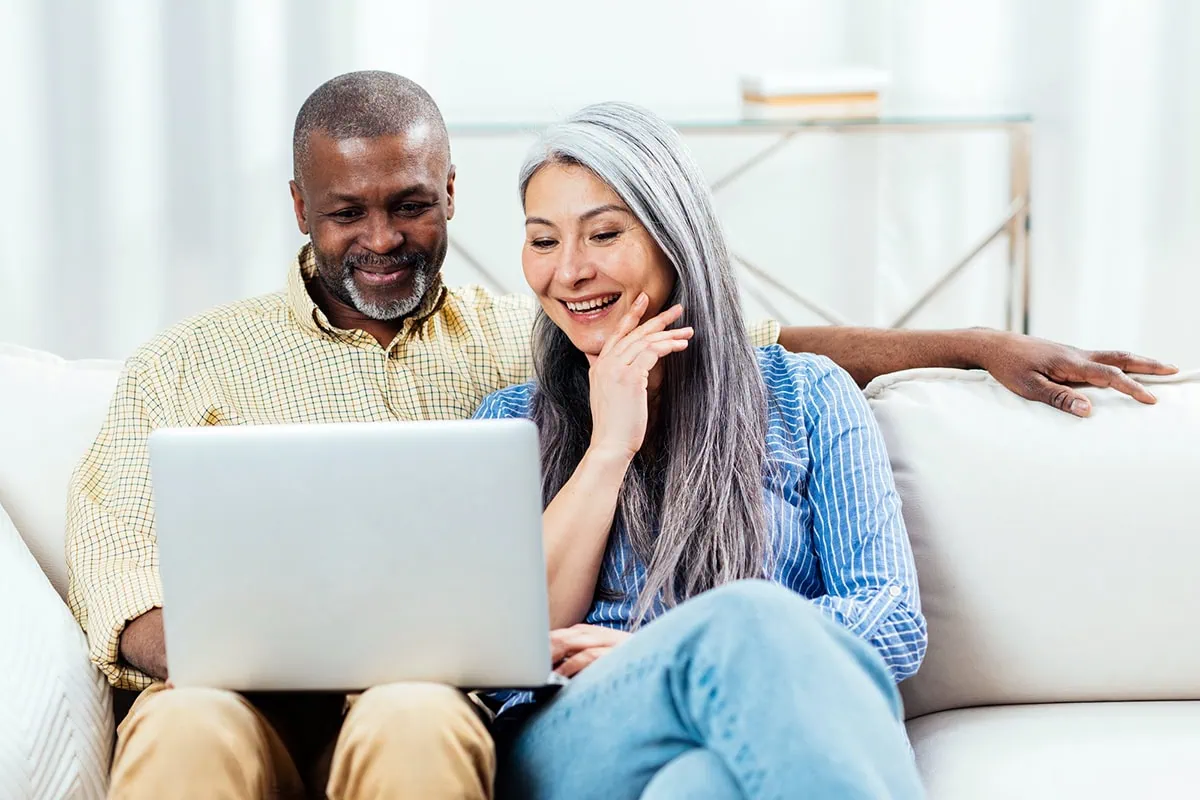a happy senior couple looks at a computer while sitting at their couch.