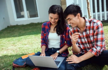 a younger couple smiles while looking at a laptop and sitting on the grass.