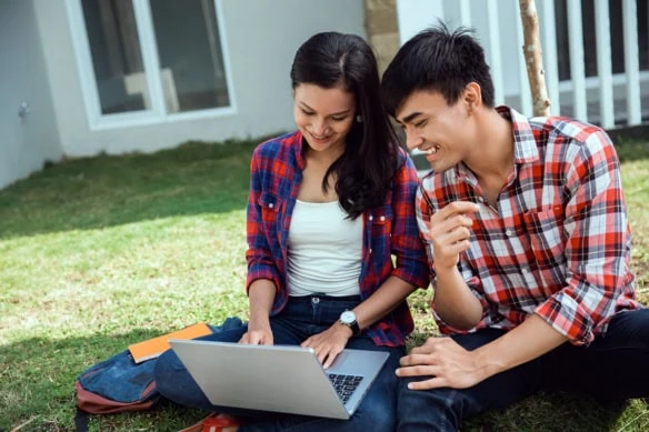 a younger couple smiles while looking at a laptop and sitting on the grass.
