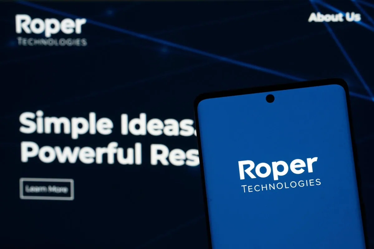 roper technologies logo on a phone held in front of a screen showing the roper technologies website.
