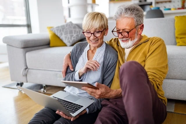 senior couple looking at laptop and tablet together beside the couch happy