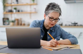 An adult woman writes in a notebook at a table with a laptop. Mature entrepreneur.