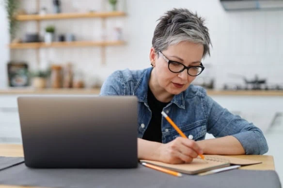 An adult woman writes in a notebook at a table with a laptop. Mature entrepreneur.