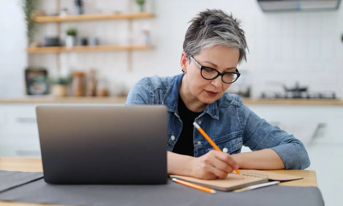 An adult woman writes in a notebook at a table with a laptop. Mature entrepreneur.